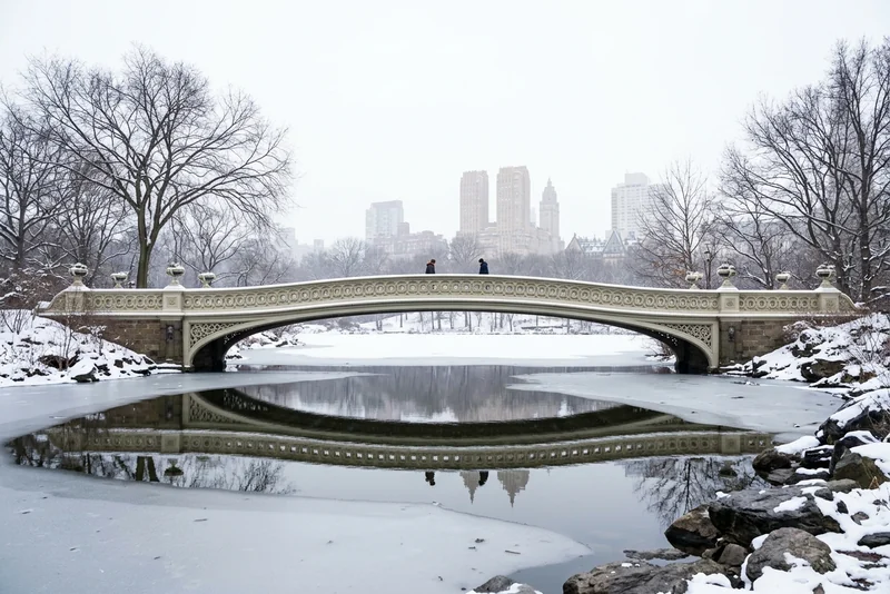 Two people crossing Bow Bridge over the frozen Lake in Central Park after snow