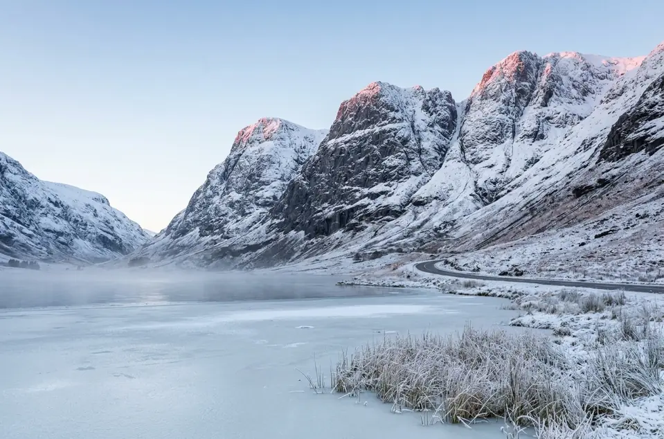 Snowy peaks lit pink at dawn above a frozen loch and the valley road in Glencoe