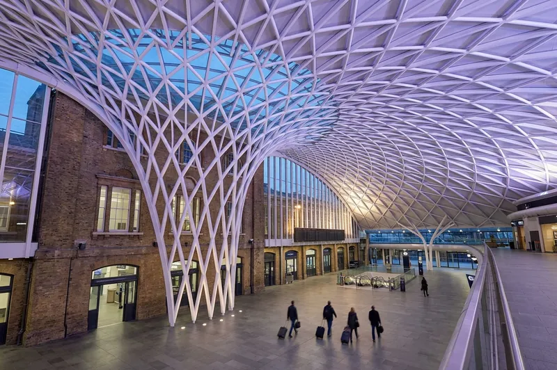 Travelers with suitcases under the white lattice roof of the King's Cross Western Concourse