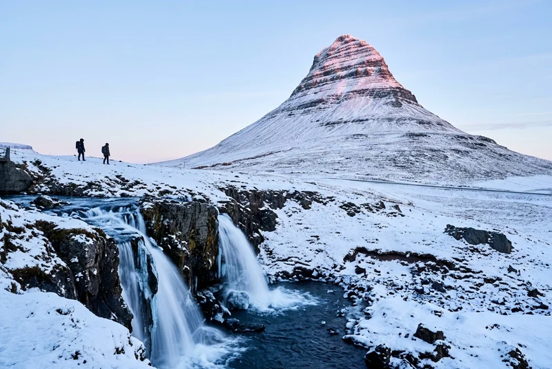 Snow-covered Kirkjufell above the half-frozen Kirkjufellsfoss falls near Grundarfjörður, two hikers on the ridge