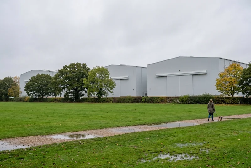 Large gray film studio buildings at Leavesden behind trees and a wet green field