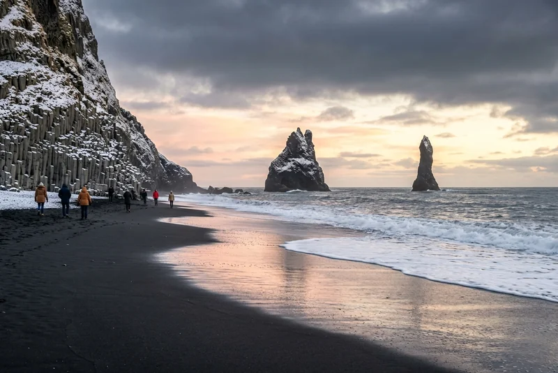 Walkers on Reynisfjara black sand beach below the basalt column cliff, with the Reynisdrangar sea stacks