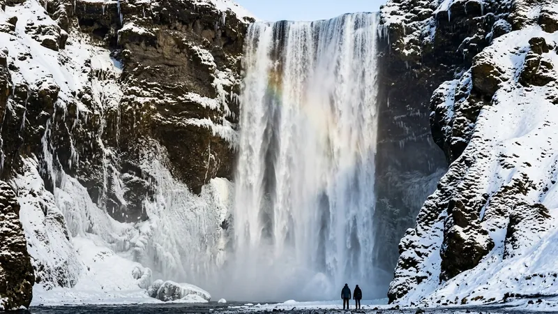 Skógafoss waterfall in winter between snowy cliffs, with a faint rainbow in the spray